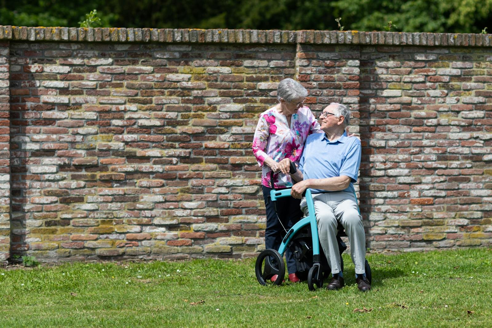 An older woman using a walking frame being supported by a companion on a footpath.